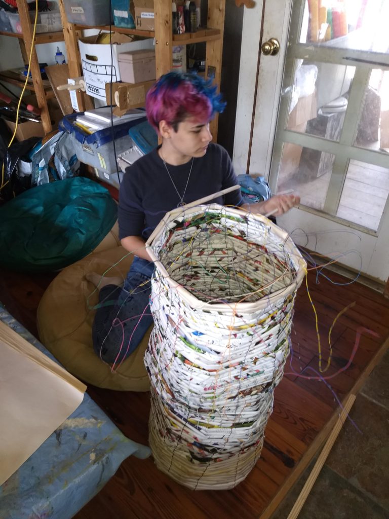 teenaged girl with purple and blue hair works on a basket that is at least 3 feet tall